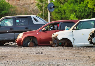 Several cars without wheels and partially stripped of parts are parked on a gravel surface, surrounded by greenery and other discarded items.